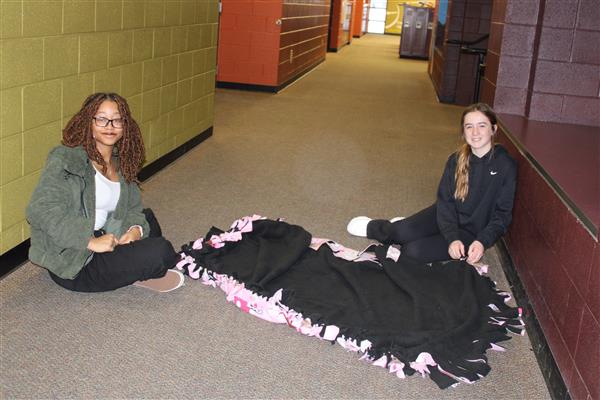 2 students sit on the ground next to a blanket that they made